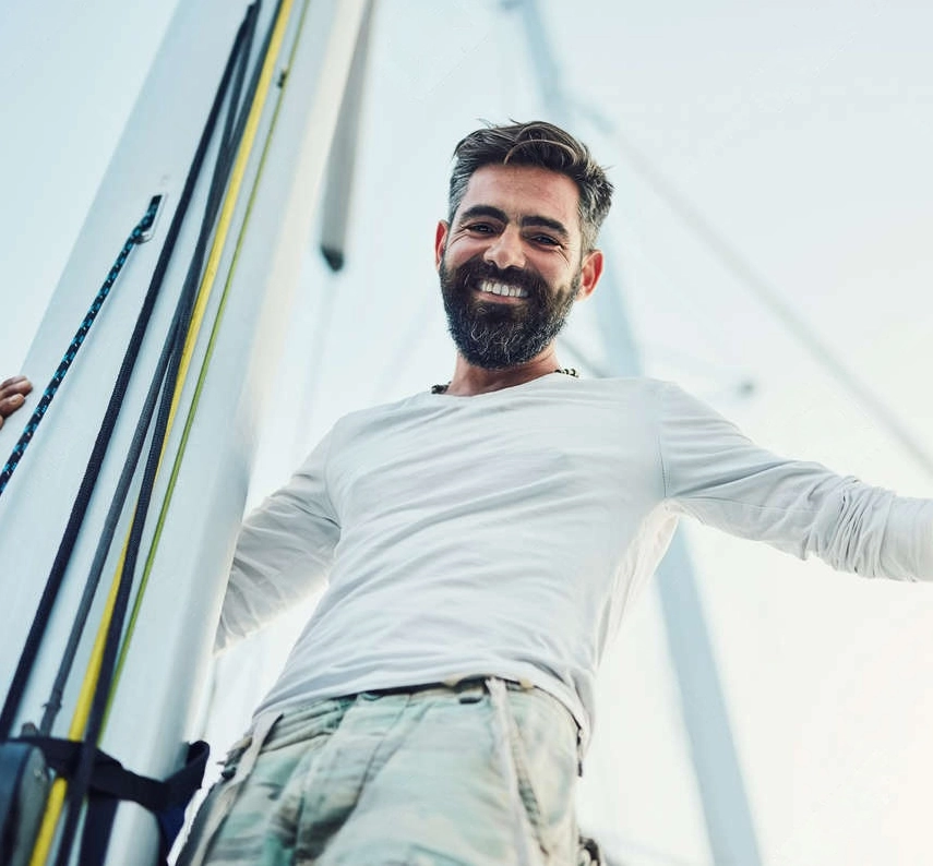 Luke Burnett in a white long-sleeve shirt stands on a sailboat, smiling, with sail lines and the mast visible in the background.