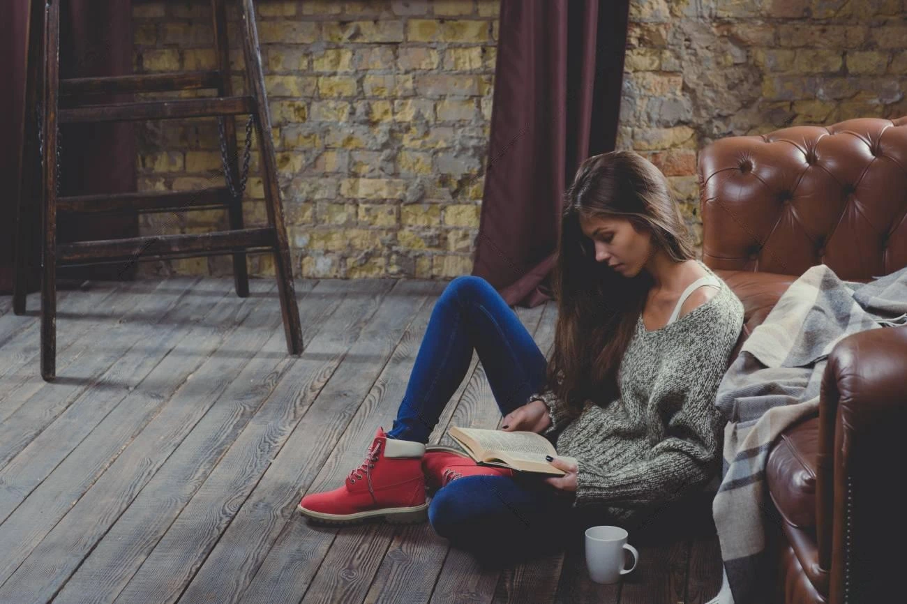 A woman sits on a wooden floor by a leather sofa, reading a book&mdash;perfect IG captions moment. She wears a grey sweater, blue jeans, and red shoes. A white mug rests on the floor next to her.