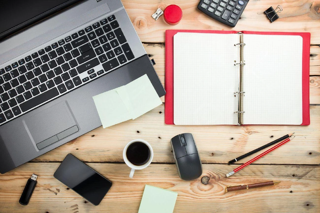 Laptop, open binder, phone, mouse, coffee, pens, sticky notes, calculator, and office supplies arranged on a wooden desk&mdash;perfect setup for productivity or crafting creative IG captions.