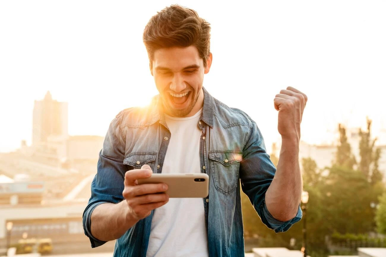 A man outdoors looks at his smartphone, smiling and raising his fist in excitement, as sunlight shines in the background&mdash;capturing the perfect moment for IG Captions.