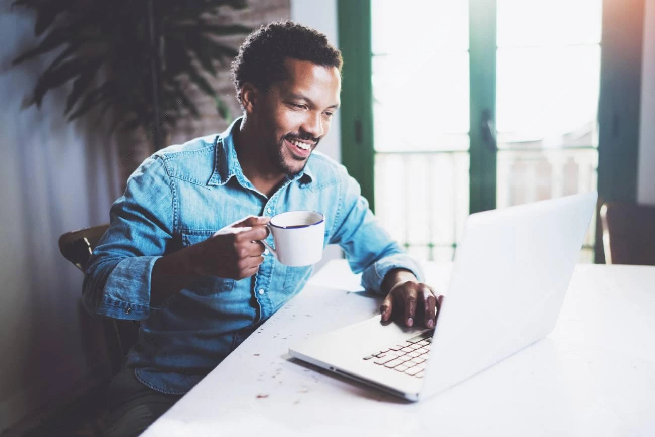 A man in a denim shirt sits at a table, holding a mug and smiling while looking at a laptop in a well-lit room&mdash;perfect moment for crafting IG captions.