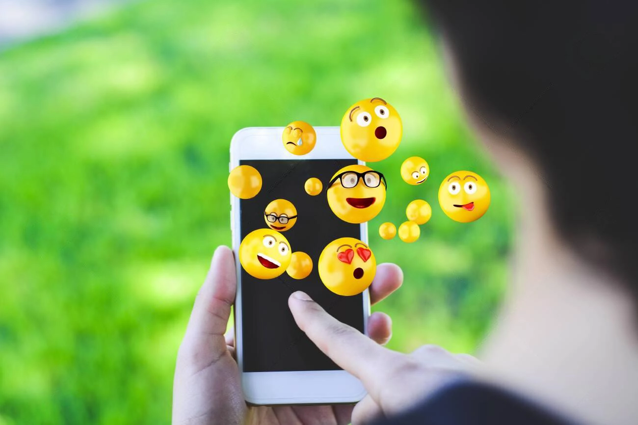 A person uses a smartphone displaying several floating emoji faces above the screen, perfect for creating fun IG Captions, with a blurred green outdoor background.