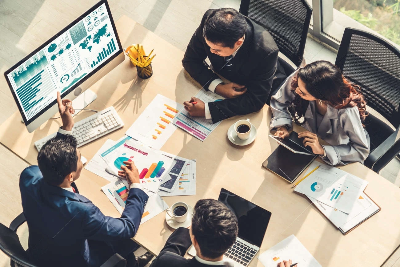 Four business people sit around a table with charts and a computer displaying graphs, discussing data and analysis in a modern office setting, while using a caption generator to enhance their presentations.