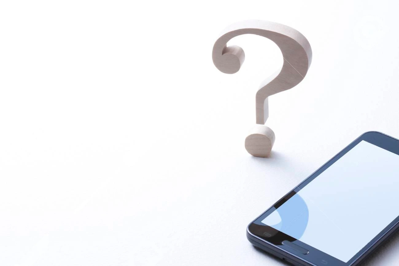 A wooden question mark stands next to a smartphone on a white surface, suggesting uncertainty or inquiry related to technology, communication, or crafting the perfect IG captions.