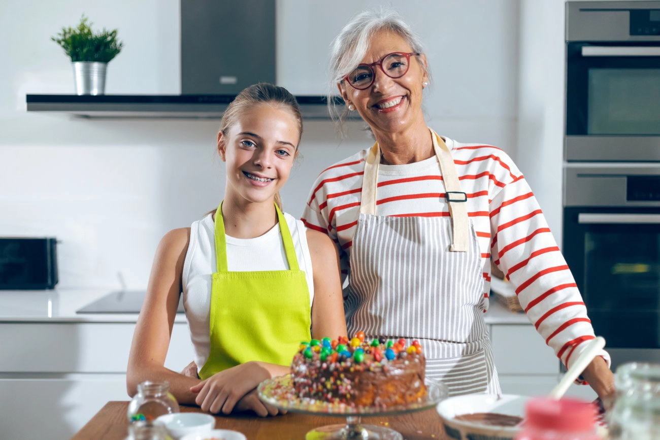 An older woman and a young girl, both wearing aprons, stand in a kitchen smiling behind a decorated chocolate cake on the counter&mdash;perfect moment for sweet IG captions.