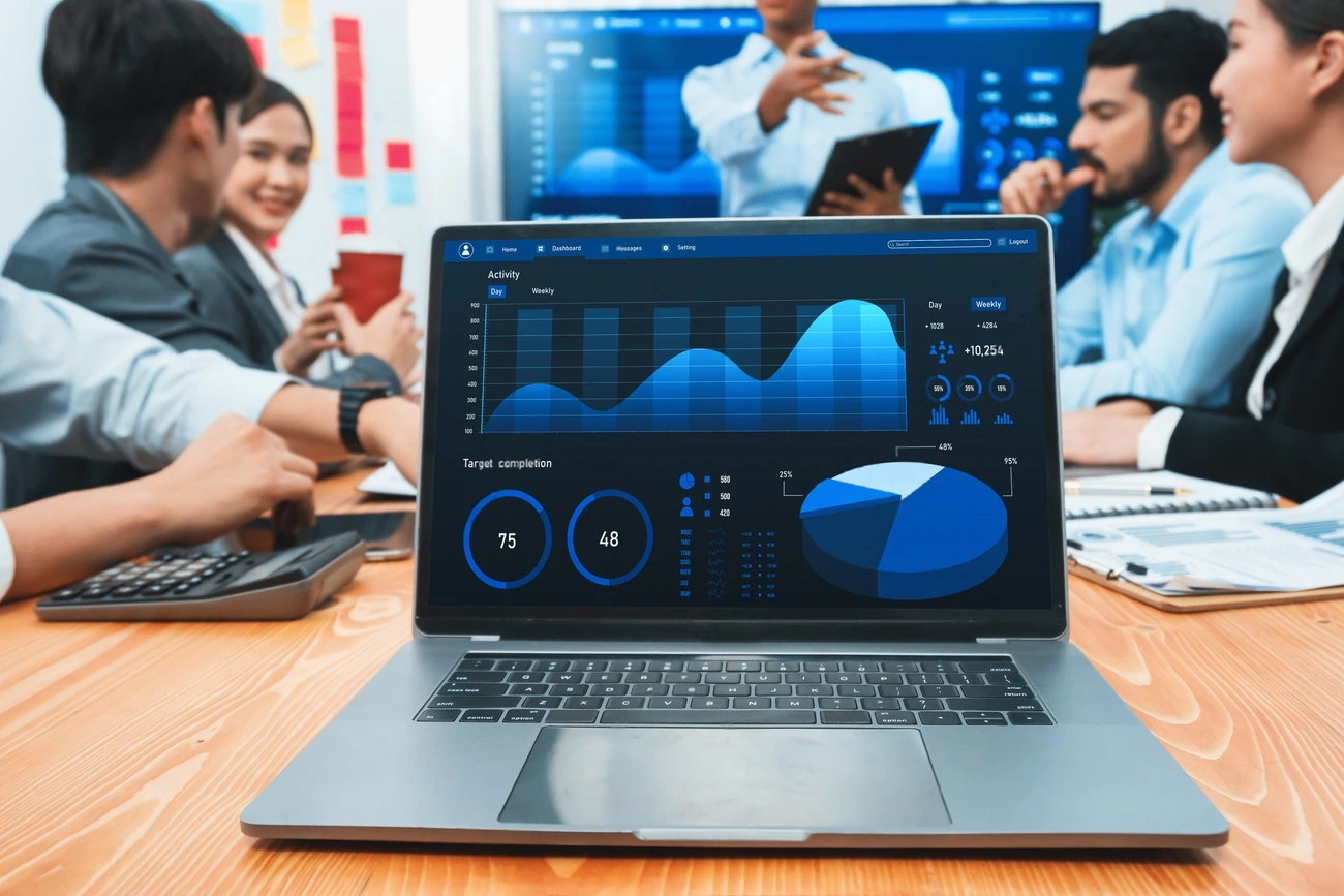 A laptop displaying graphs and data dashboards sits on a table during a business meeting, as colleagues discuss Instagram Story Views in the background.