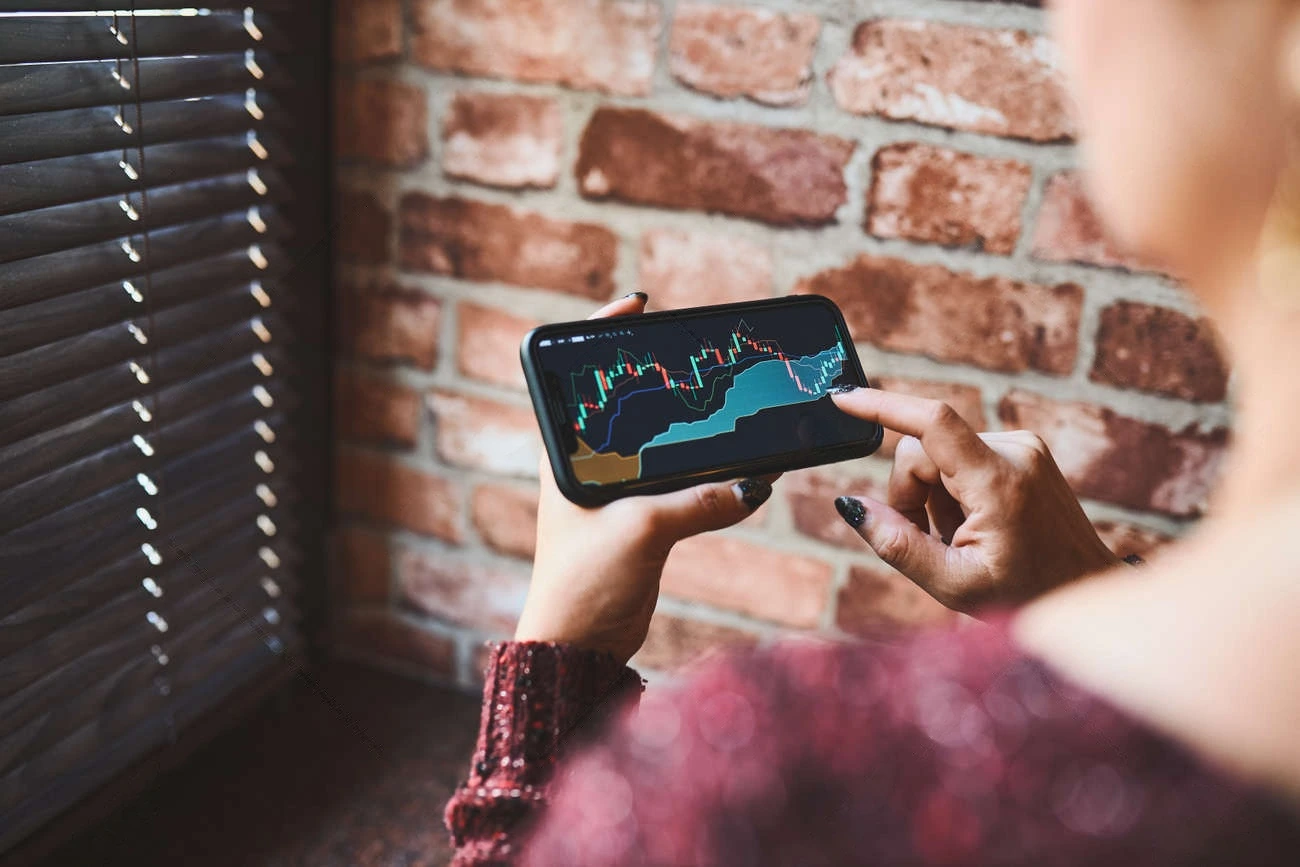 A person holds a smartphone displaying a financial chart with candlestick and area graphs, tracking Instagram Story Views, while standing near a window with blinds and a brick wall in the background.