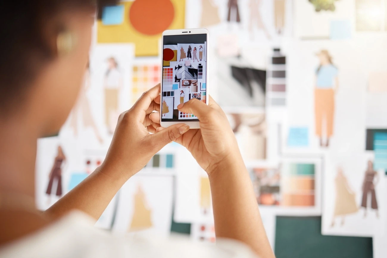 A person holds a smartphone, capturing an Instagram Story of a mood board filled with fashion sketches, color swatches, and fabric samples pinned to the wall—perfect for boosting Instagram Story Views.