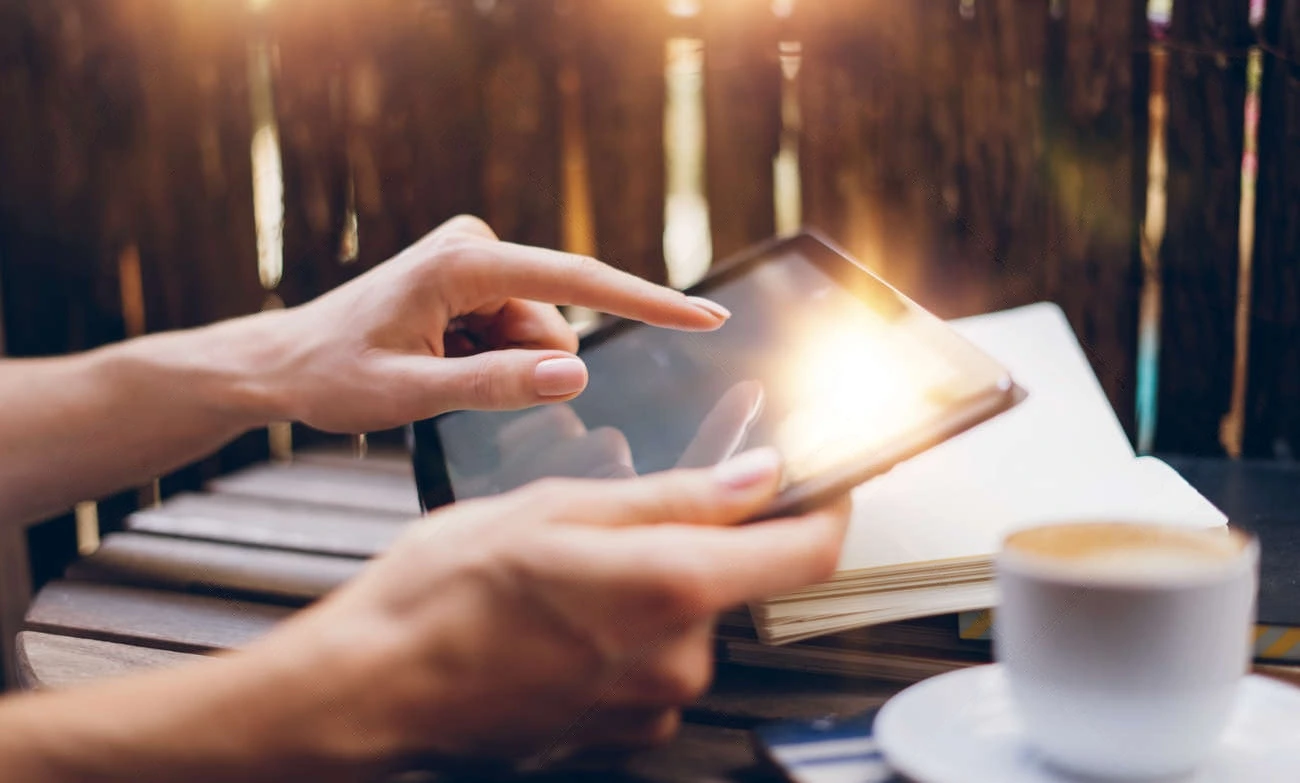 A person uses a tablet outdoors next to an open notebook and a cup of coffee on a table, possibly trying out a caption generator for their notes.