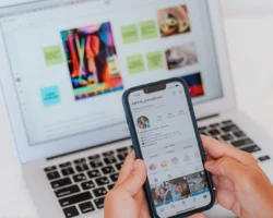 A person holds a smartphone displaying an Instagram profile in front of a laptop with colorful notes and images on its screen, possibly brainstorming creative IG Captions.