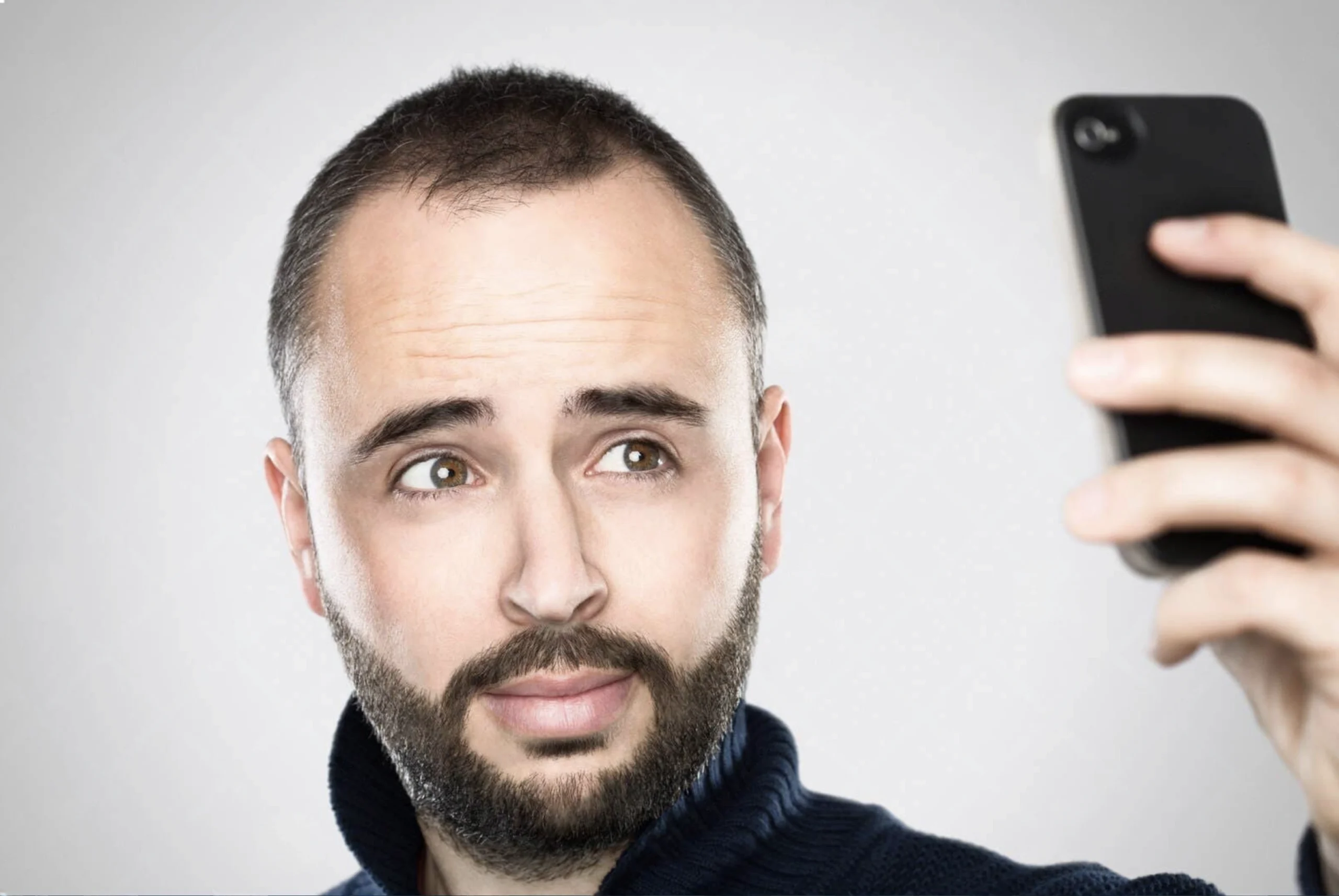 A man with a short beard and receding hairline looks slightly to the side while holding a smartphone up to take a selfie, perfect for a candid PFP.