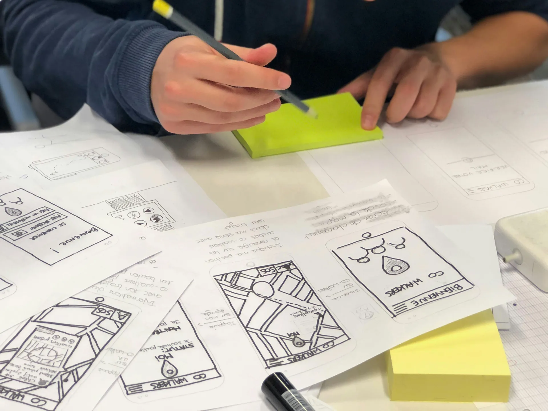 A person sketches mobile app wireframes with a pencil on paper and sticky notes, surrounded by several design drafts and documents.