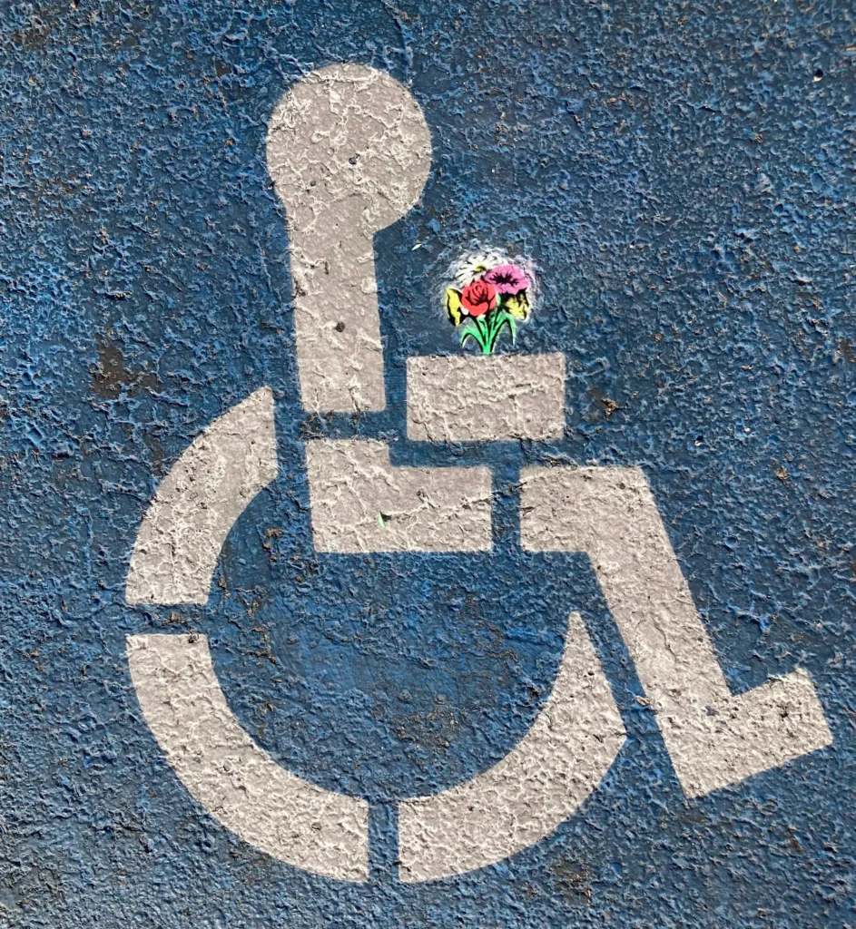 A white wheelchair accessibility symbol on blue pavement, with a small colorful flower drawn on the armrest area, highlights the importance of digital accessibility in creating inclusive environments for all.